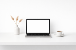 ​Laptop sobre escritorio minimalista blanco con taza de café y elementos decorativos, espacio de trabajo moderno.